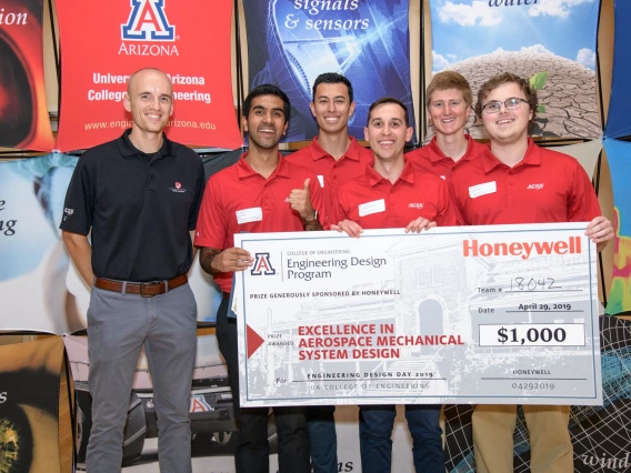 A group of five students in red shirts holding a giant check. Their project sponsor stands next to them in a blue shirt.