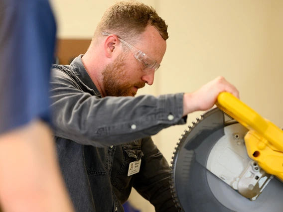 A man with red hair cuts a piece of metal with a large saw.