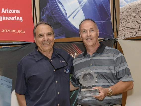 Two men in short-sleeved shirts pose for a photo. The older gentleman on the right is holding a glass award.