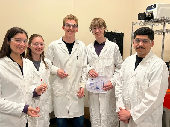 A group of young people pose for a photo in lab coats, holding a catheter system.