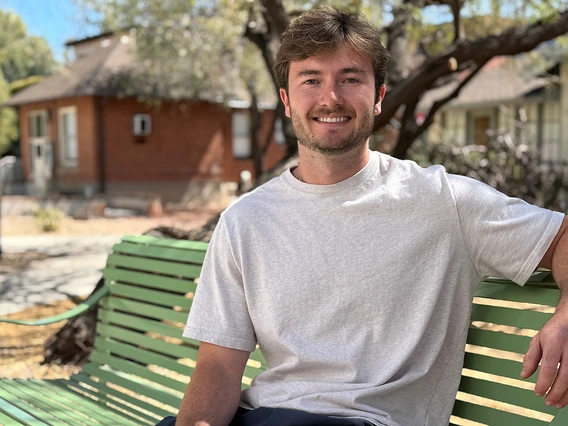 A young man sits on a bench in front of green trees.