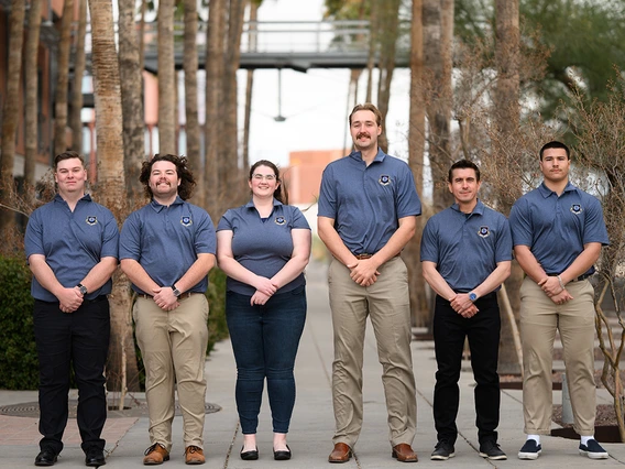 Six people in grey blue shirts pose for a photo in front of palm trees.