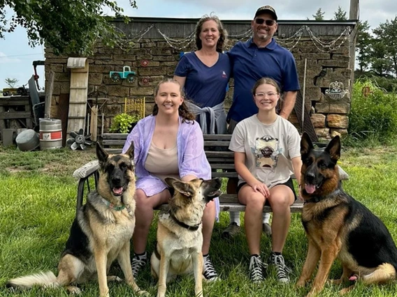 A man in a blue shirt takes a photo with his wife and two daughters. They also have three German shepherds.
