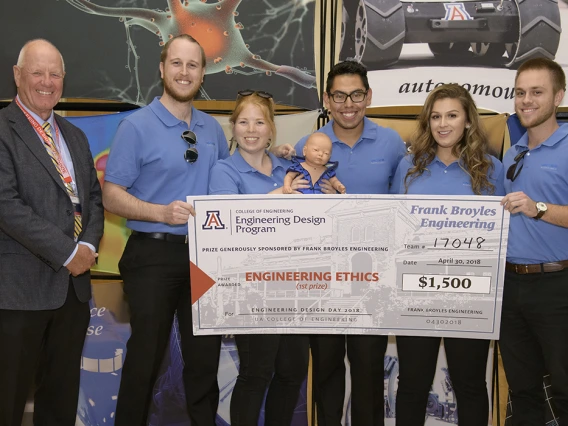 A man in a suit standing next to five students wearing blue polo shirts and holding a giant check for $1,500