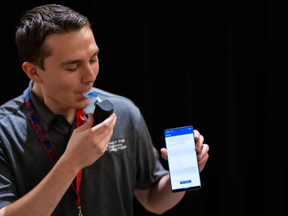 A student wearing a button up shirt holds a phone while demonstrating how to use a breathing device.