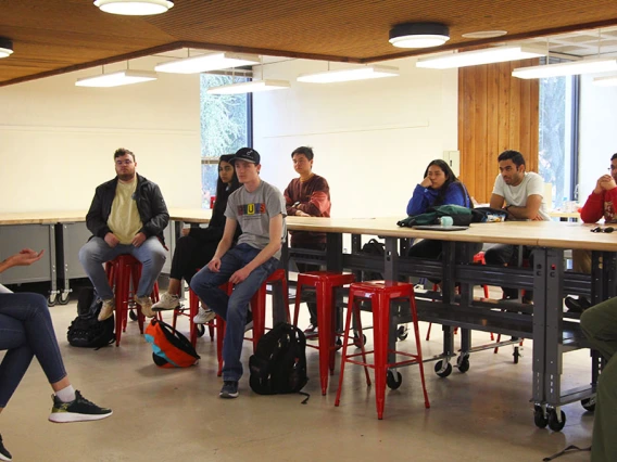 One student addresses a group of others in a room with tables and stools.