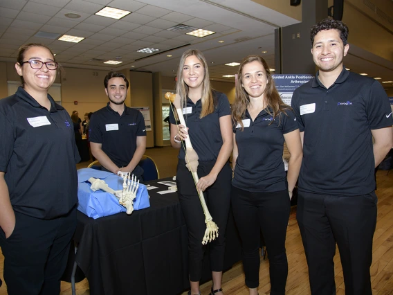 Five students on an engineering design team standing in a ballroom holding a skeletal leg with another on the table