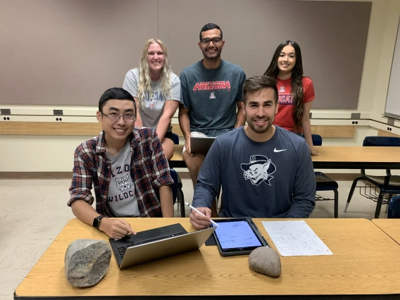 Five students in a classroom. Two sit at a table with a laptop in front of them, and three are standing behind them.