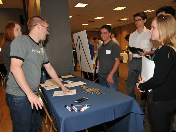 Two people behind a table speak to four university students on the other side of the table at an open house