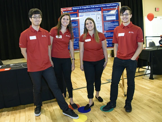 Four university students in red shirts standing in front of a presentation board