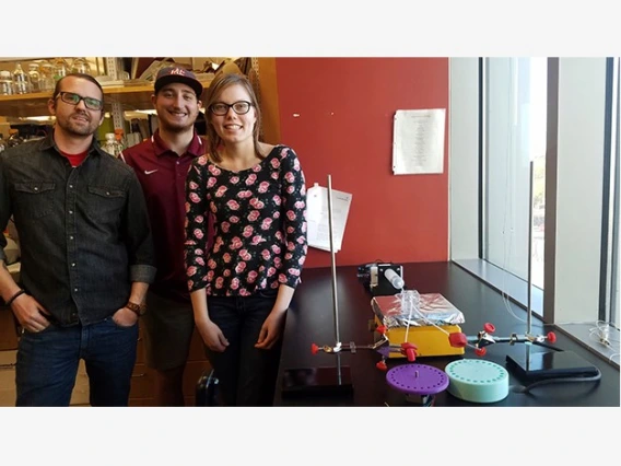 Three university students standing next to an apparatus they designed to mimic features of the human eye