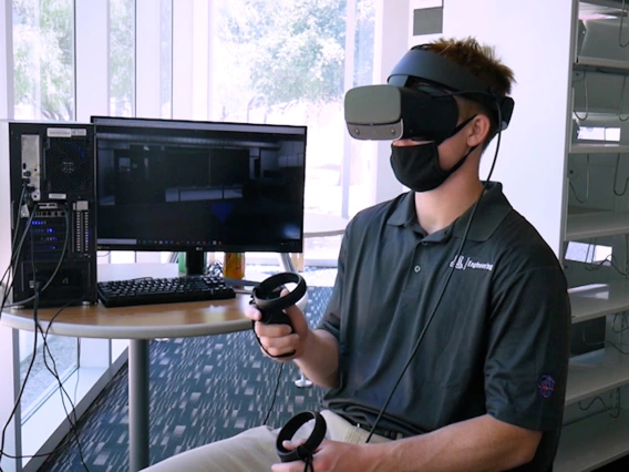 A student wearing a virtual reality headset and sitting next to a computer monitor.