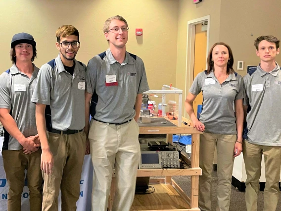 a group of 7 people stands with a cart displaying materials used for an engineering project