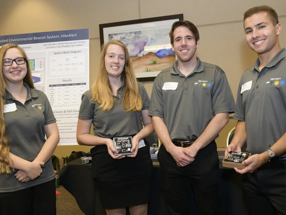 Four students in gray polo shirts stand in front of a scientific poster, holding electronic parts of a beacon system.