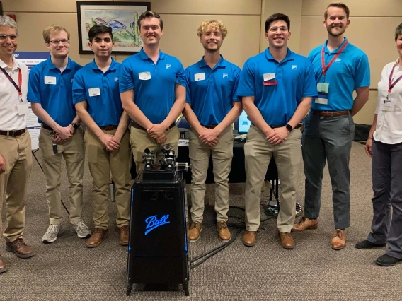 8 people pose indoors with a waist-high piece of equipment