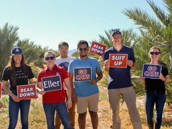 Eight University of Arizona students holding school signs in a date palm field