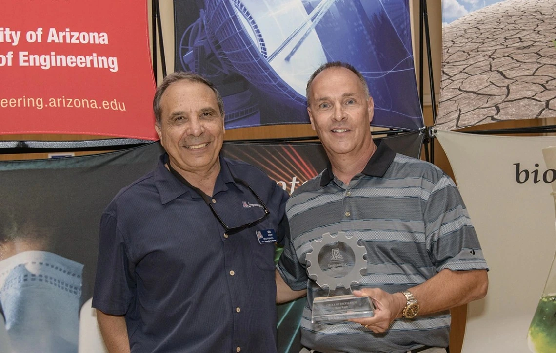 Two men in short-sleeved shirts pose for a photo. The older gentleman on the right is holding a glass award.