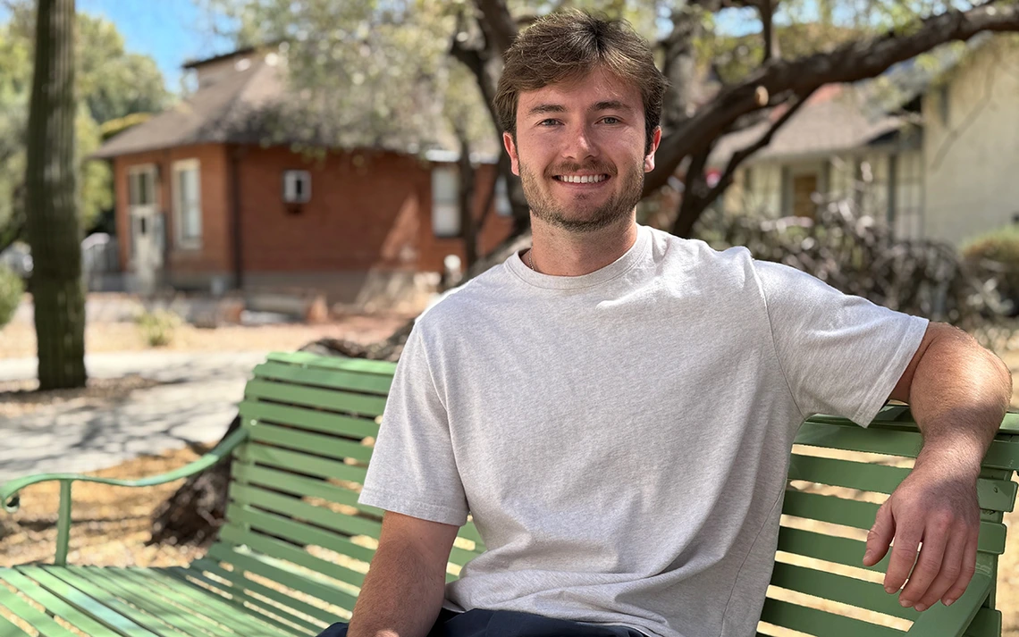 A young man sits on a bench in front of green trees.