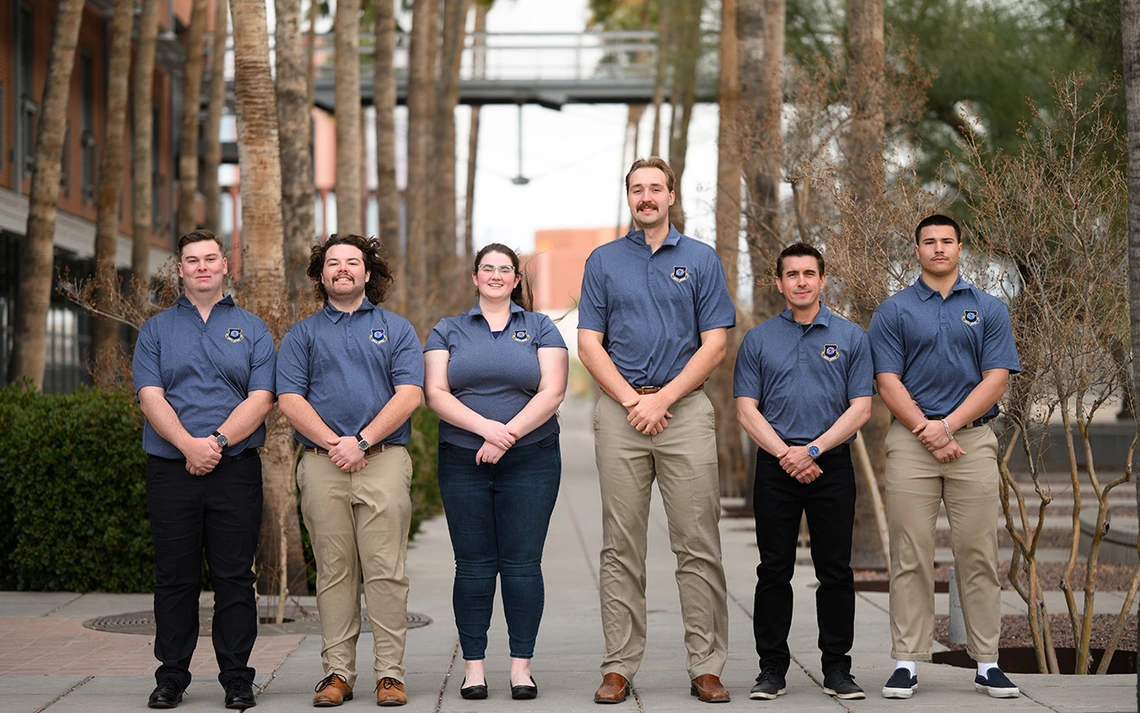 Six people in grey blue shirts pose for a photo in front of palm trees.