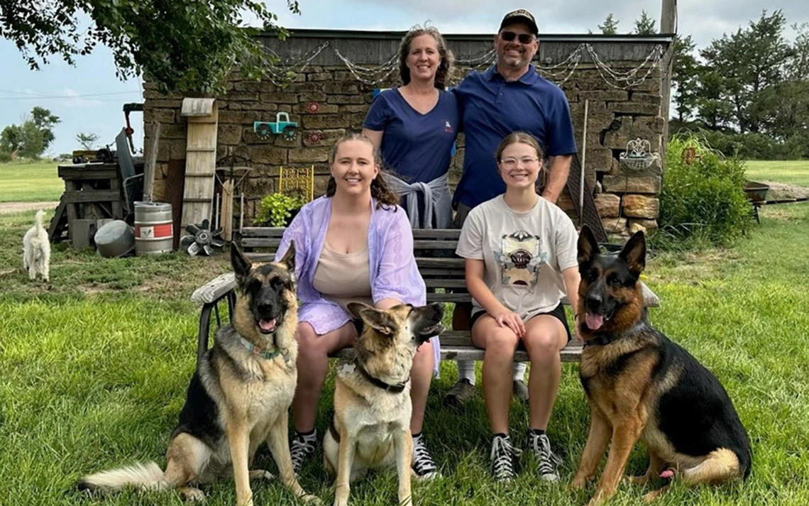 A man in a blue shirt takes a photo with his wife and two daughters. They also have three German shepherds.