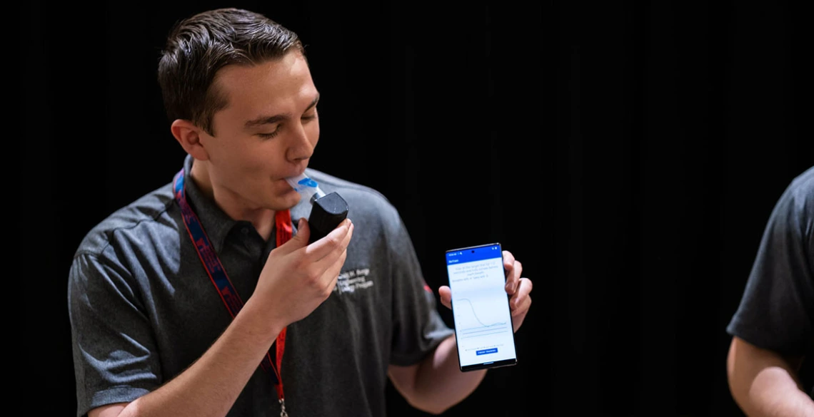 A student wearing a button up shirt holds a phone while demonstrating how to use a breathing device.