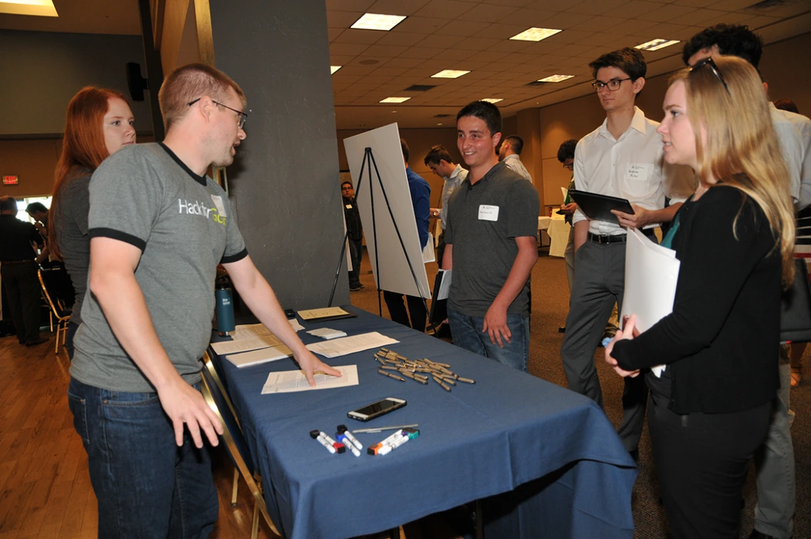 Two people behind a table speak to four university students on the other side of the table at an open house