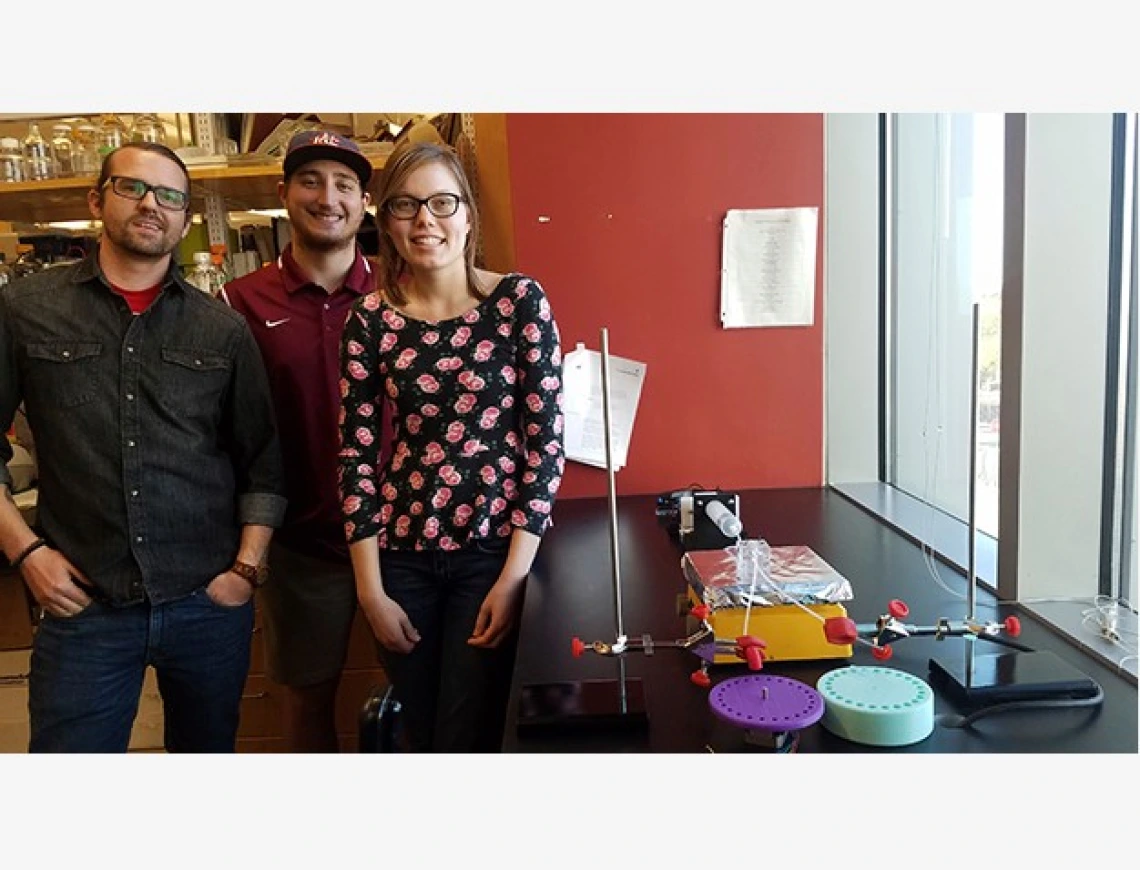 Three university students standing next to an apparatus they designed to mimic features of the human eye