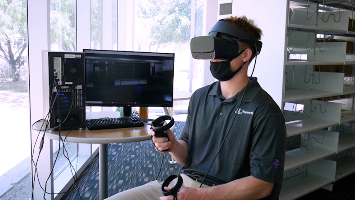 A student wearing a virtual reality headset and sitting next to a computer monitor.