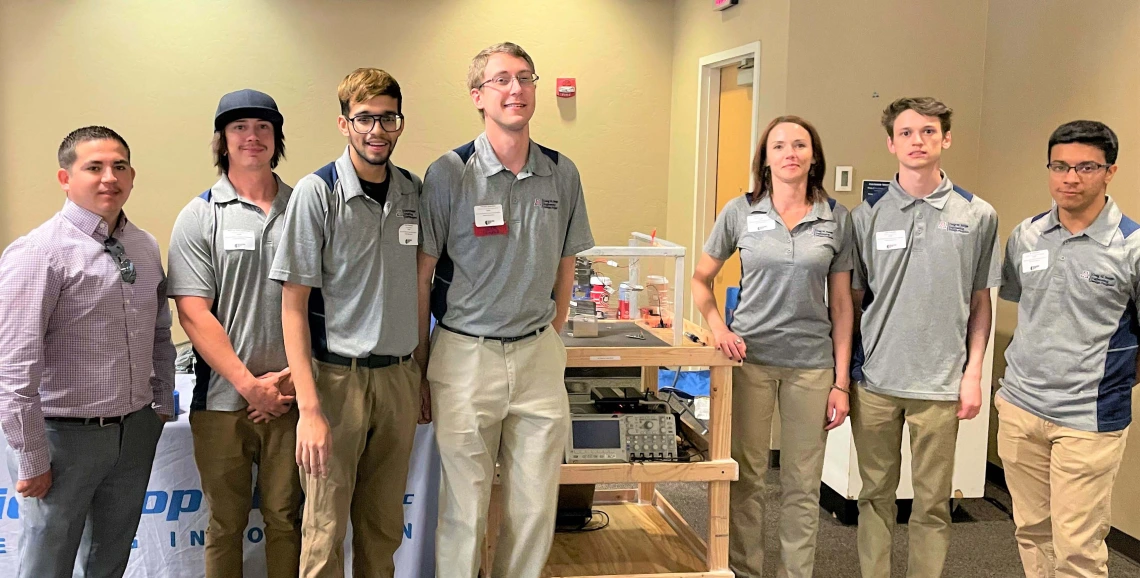 a group of 7 people stands with a cart displaying materials used for an engineering project