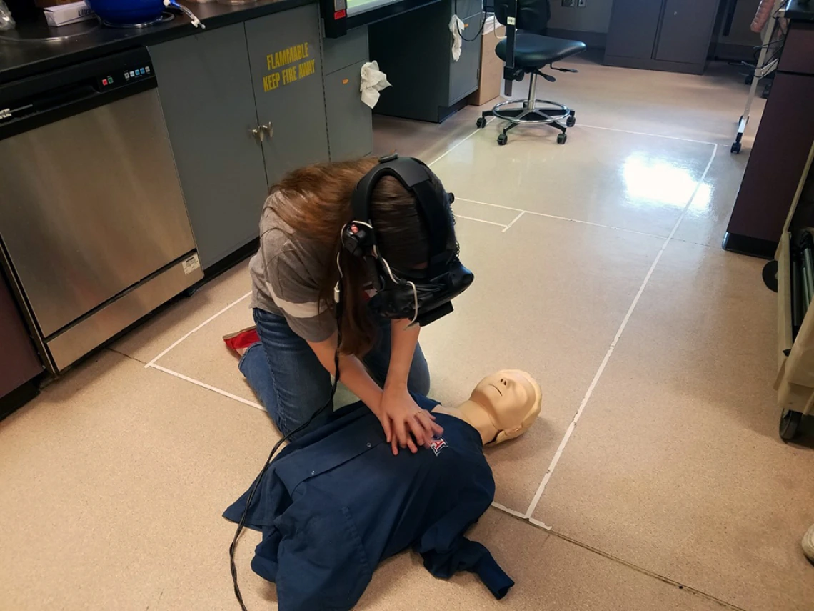 A woman wearing a virtual reality headset kneeling over a CPR practice dummy.