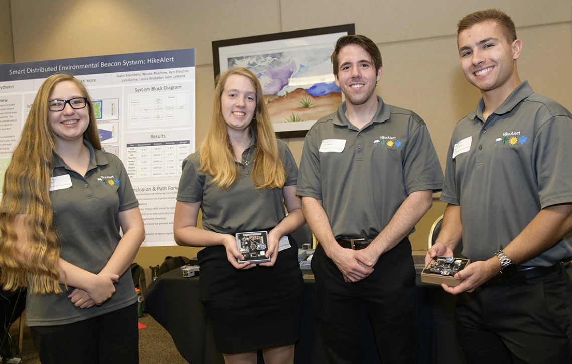 Four students in gray polo shirts stand in front of a scientific poster, holding electronic parts of a beacon system.