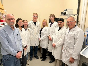 Five students and two doctors in lab clothes stand in a lab with an older man in a blue long-sleeve shirt.