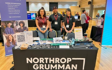 Three people stand behind a booth labeled "Northrup Grumman" and pose for a picture at a student fair.