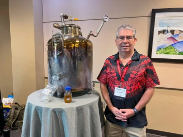 A man in a red and black shirt stands next to a pyrolysis devices and a sample of the fuel created with the device.