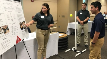 A female students explains information on a poster board to two male students