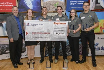 A team of five students in gray polo shirts hold a comically large check, while a man in a gray blazer and blue shirt stands next to them smiling. Several clear trophies are resting on the floor in front of them.