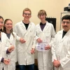 A group of young people pose for a photo in lab coats, holding a catheter system.