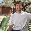 A young man sits on a bench in front of green trees.