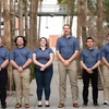 Six people in grey blue shirts pose for a photo in front of palm trees.