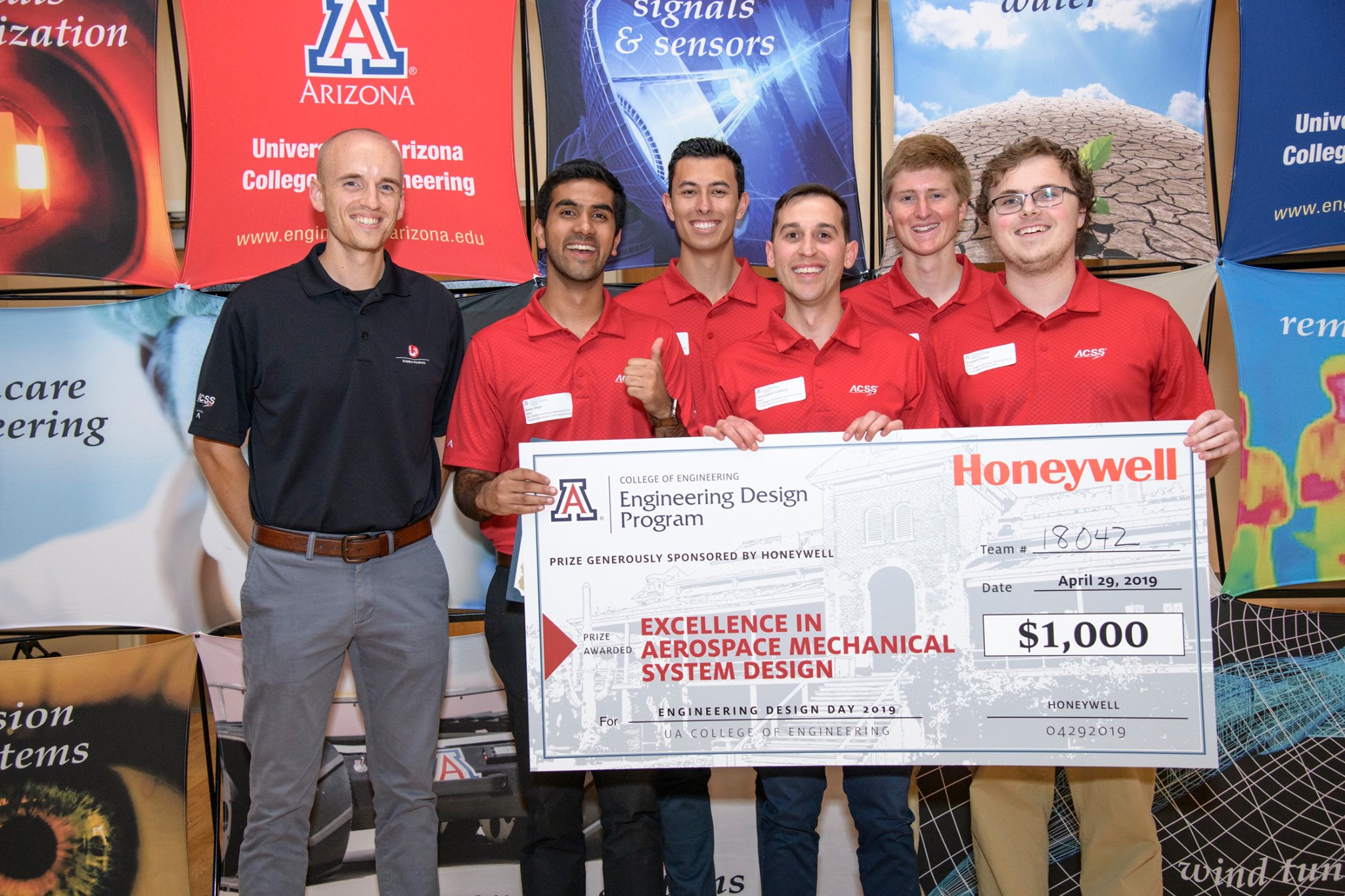 A group of five students in red shirts holding a giant check. Their project sponsor stands next to them in a blue shirt.
