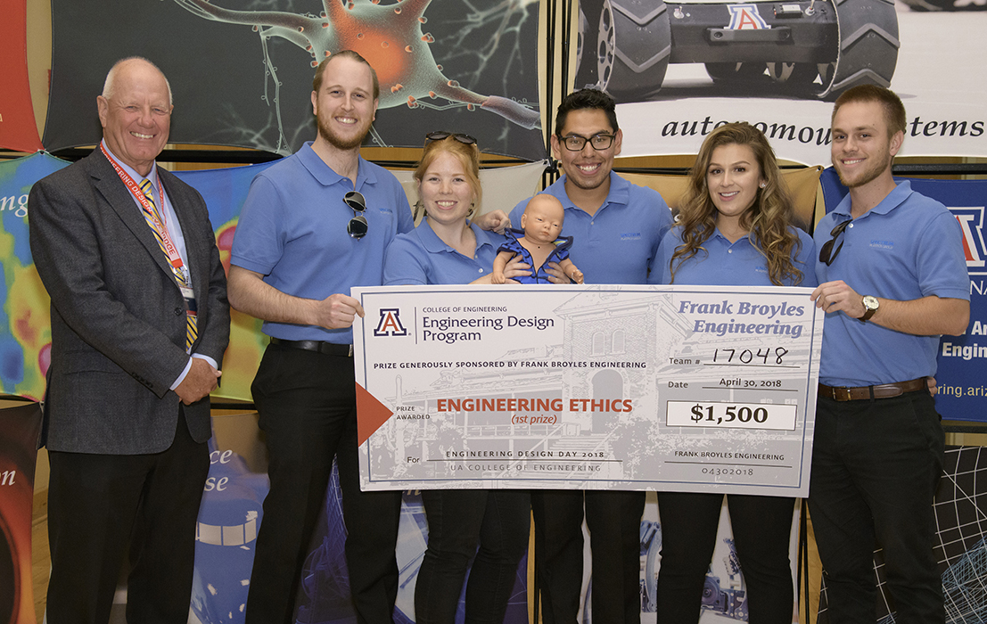 A man in a suit standing next to five students wearing blue polo shirts and holding a giant check for $1,500