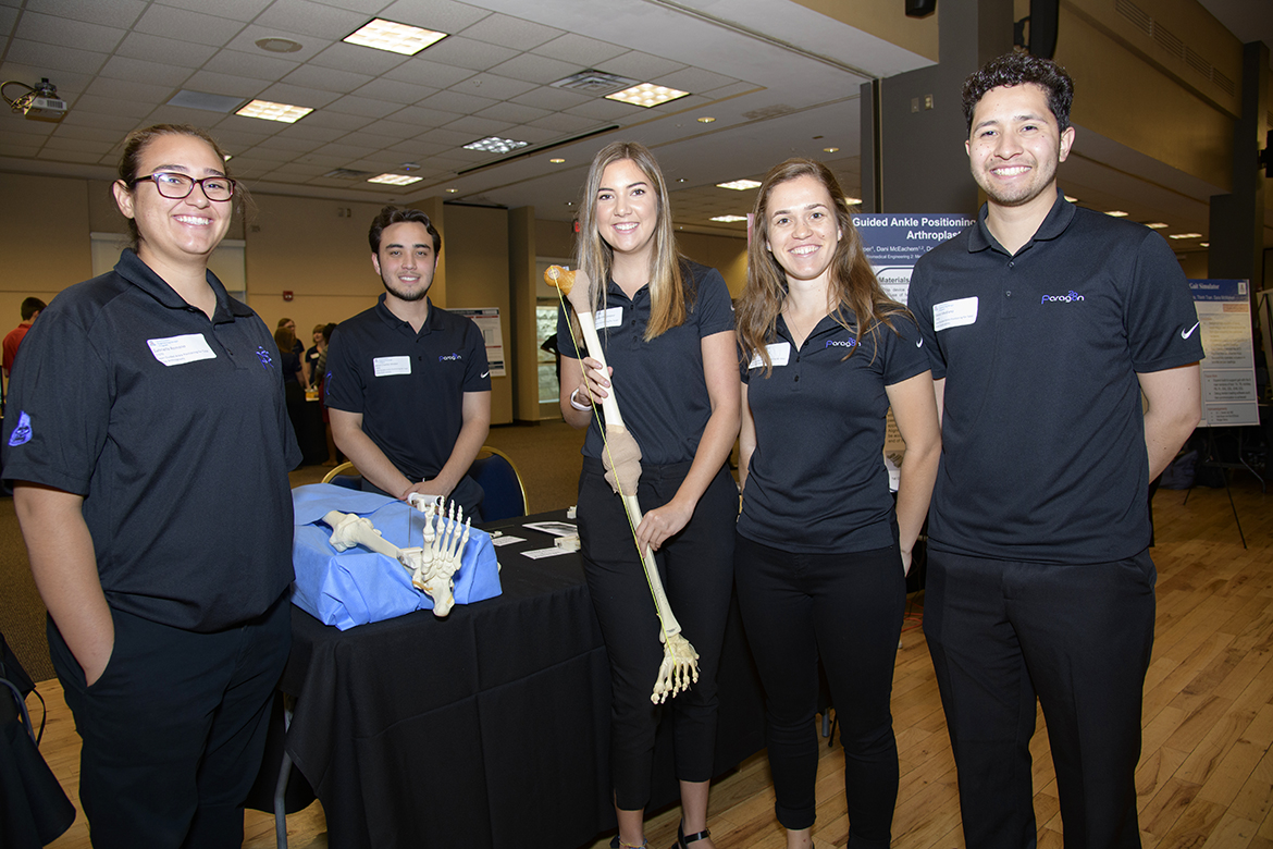 Five students on an engineering design team standing in a ballroom holding a skeletal leg with another on the table