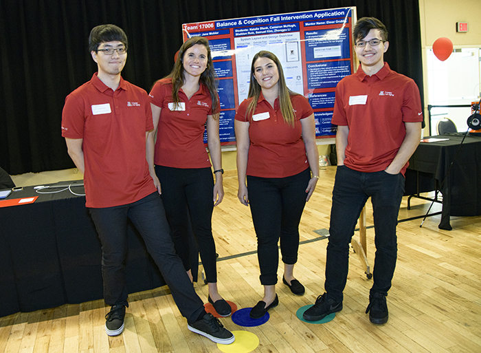 Four university students in red shirts standing in front of a presentation board