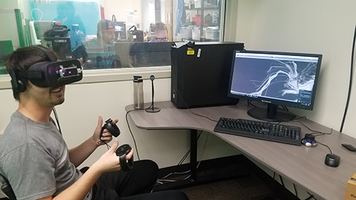 A student wearing VR goggles and handling two control pads looks around while a nearby monitor displays a computer-generated representation of a brain's white matter fiber tracts