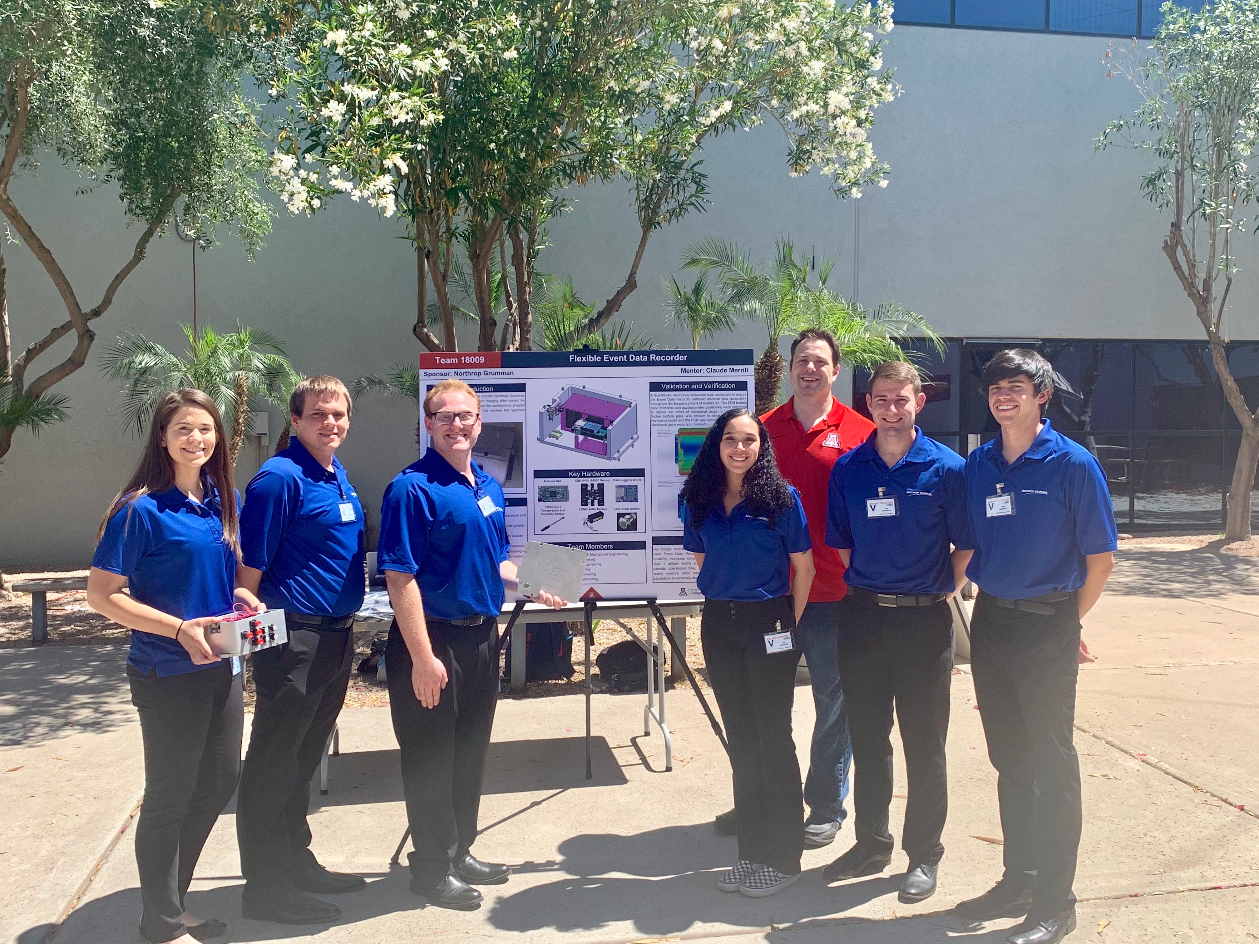 A group of seven people, six in blue polos and one in a red polo) stand next to an academic poster. They are outside and a tree is in the background.