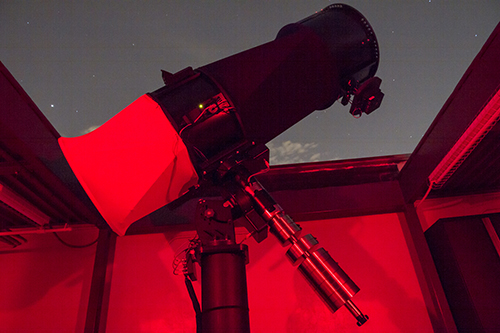 Photo of a telescope pointing at the night sky out of an opening in a roof with a red light on inside
