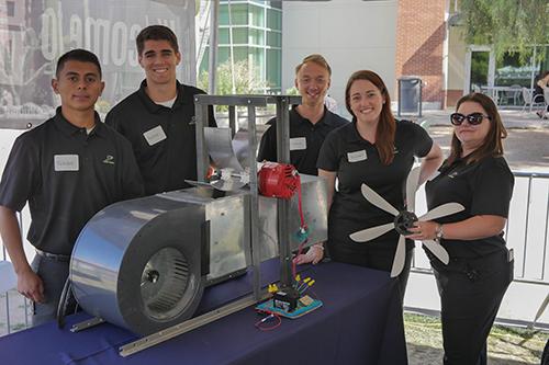 Three male and two female university students under a tent oudoors, standing by a table holding a wind turbine model