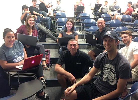 A group of seven university students in a classroom setting, four of which have laptops