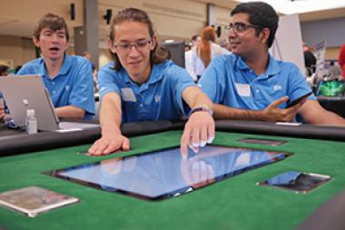 Three university students in blue shirts around a touchscreen display embedded in a table