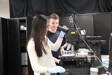 A female member of Team 17047 selects a slide while her other hand rests on an electron microscope while a male team member observes